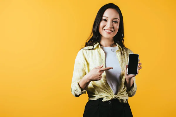 Lächeln asiatisches Mädchen zeigt auf Smartphone mit leerem Bildschirm isoliert auf gelb — Stockfoto