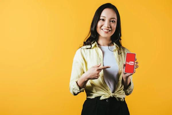 KYIV, UCRANIA - 16 DE ABRIL DE 2019: atractiva chica asiática sonriente apuntando al teléfono inteligente con la aplicación de youtube, aislada en amarillo - foto de stock