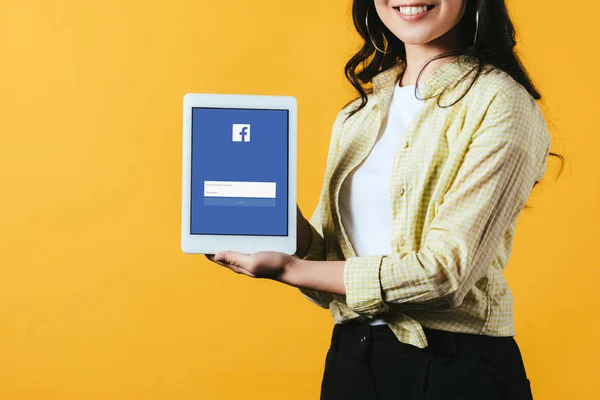 KYIV, UKRAINE - APRIL 16, 2019: cropped view of smiling girl showing digital tablet with facebook app, isolated on yellow — Stock Photo