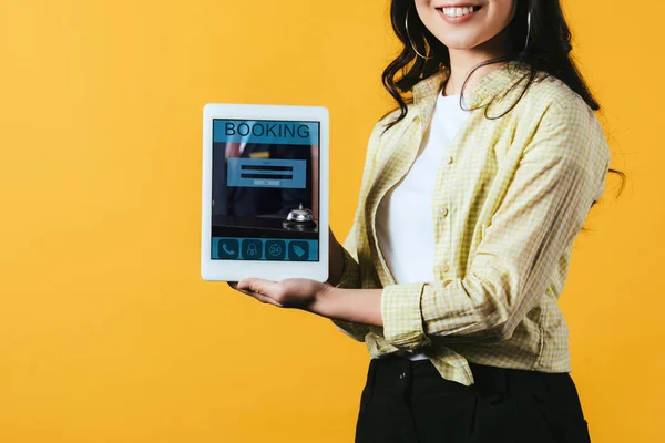 Обрезанный вид девушки, показывающей цифровой планшет с приложением бронирования, изолированный на желтый — стоковое фото