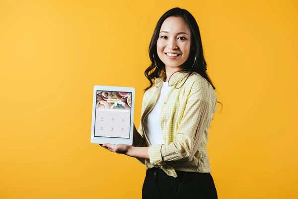 KYIV, UCRANIA - 16 DE ABRIL DE 2019: chica asiática sonriente mostrando tableta digital con aplicación cuadrada, aislada en amarillo - foto de stock