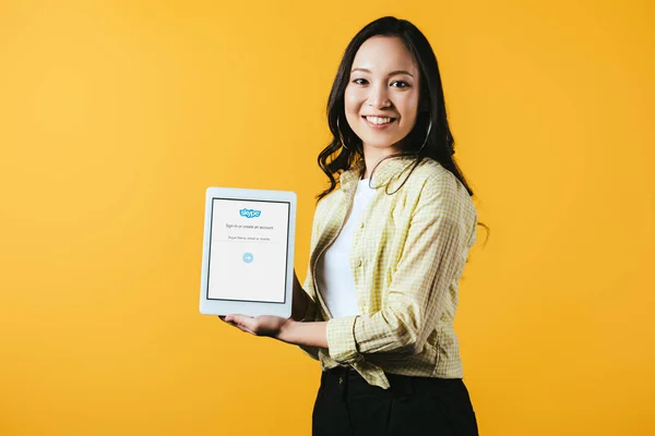 KYIV, UCRANIA - 16 DE ABRIL DE 2019: hermosa chica asiática mostrando tableta digital con aplicación skype, aislada en amarillo - foto de stock