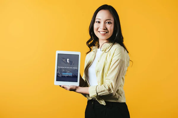 KYIV, UCRANIA - 16 DE ABRIL DE 2019: hermosa chica asiática mostrando tableta digital con aplicación tumblr, aislada en amarillo - foto de stock