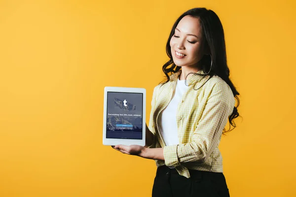KYIV, UCRANIA - 16 DE ABRIL DE 2019: chica asiática sonriente mostrando tableta digital con aplicación tumblr, aislada en amarillo - foto de stock