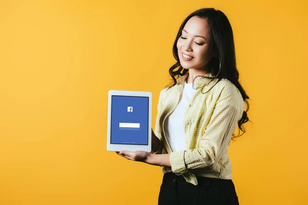 KYIV, UCRANIA - 16 DE ABRIL DE 2019: hermosa chica asiática mostrando tableta digital con aplicación de facebook, aislado en amarillo - foto de stock