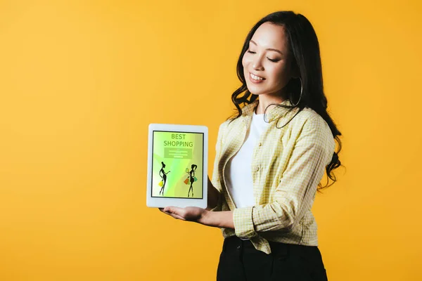 Счастливая азиатская девушка показывает цифровой планшет с лучшим приложением для покупок, изолированный на желтый — стоковое фото