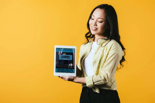 Feliz chica asiática mostrando tableta digital con aplicación de reserva, aislado en amarillo - foto de stock