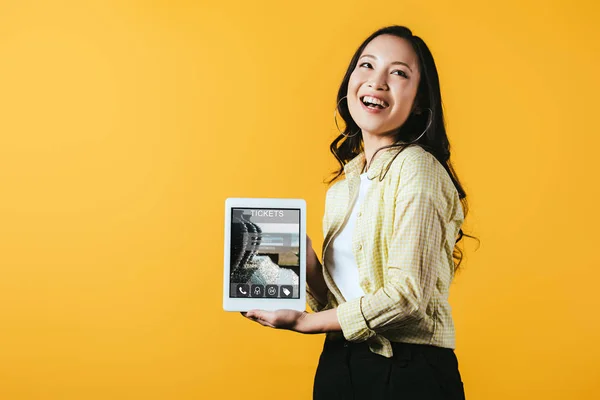 Smiling asian girl showing digital tablet with tickets app, isolated on yellow — Stock Photo