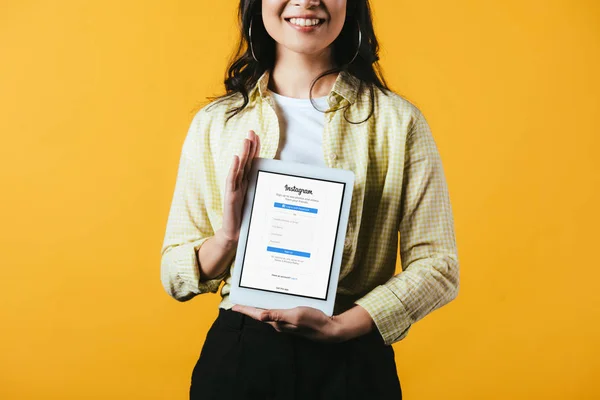 KYIV, UCRANIA - 16 DE ABRIL DE 2019: vista recortada de una chica sonriente mostrando una tableta digital con aplicación instagram, aislada en amarillo - foto de stock