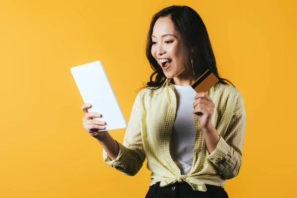 Збуджена азіатська дівчина купує онлайн з цифровим планшетом і кредитною карткою, ізольована на жовтому — стокове фото