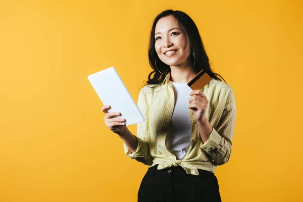 Chica asiática soñadora compras en línea con tableta digital y tarjeta de crédito, aislado en amarillo - foto de stock