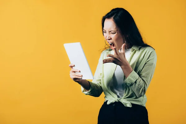 Сердитая азиатка кричит и использует цифровой планшет, изолированный на желтый — стоковое фото