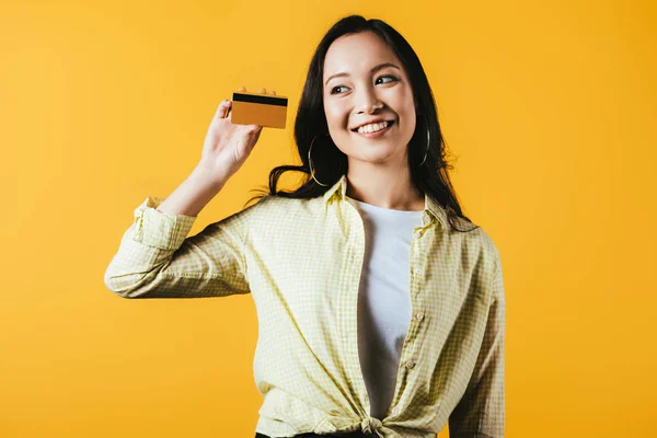 Alegre asiático menina segurando cartão de crédito, isolado no amarelo — Fotografia de Stock