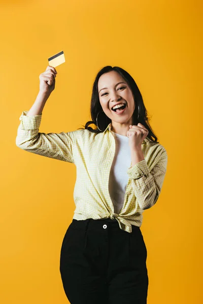 Excitada chica asiática sosteniendo tarjeta de crédito, aislado en amarillo - foto de stock