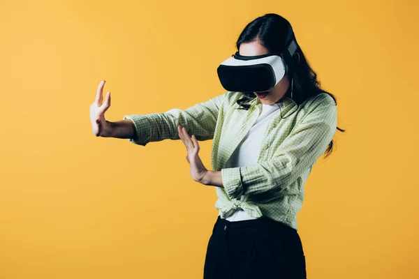 Mujer joven haciendo gestos en auriculares de realidad virtual, aislados en amarillo - foto de stock