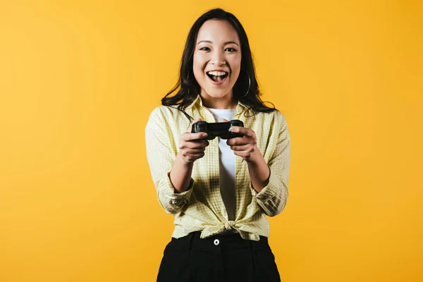 KYIV, UKRAINE - 16 AVRIL 2019 : femme asiatique excitée jouant à un jeu vidéo avec joystick, isolée sur jaune — Photo de stock