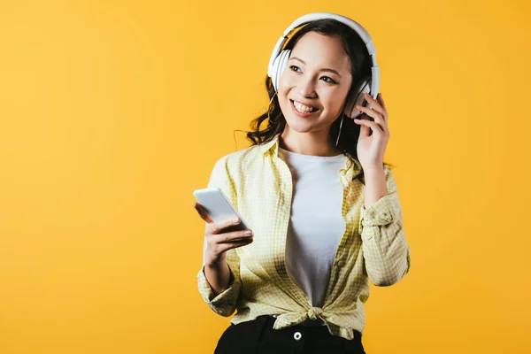 Улыбающаяся азиатская девушка, слушающая музыку с наушниками и смартфоном, изолированная на желтом — стоковое фото