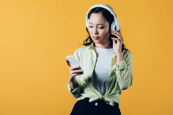 Asian girl listening music with headphones and smartphone, isolated on yellow — Stock Photo