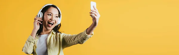 Happy asian girl taking selfie on smartphone, singing and listening music with headphones, isolated on yellow — Stock Photo