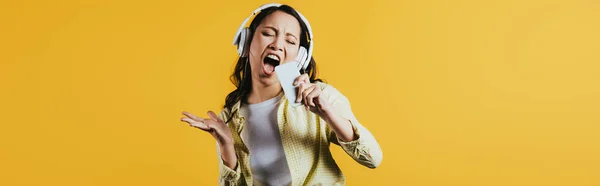 Емоційна азіатська дівчина співає і слухає музику з навушниками та смартфонами, ізольована на жовтому — стокове фото