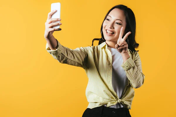 Attractive asian girl showing peace symbol and taking selfie on smartphone isolated on yellow — Stock Photo