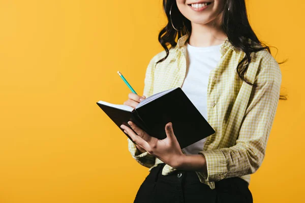 Abgeschnittene Ansicht eines lächelnden Mädchens, das mit Stift in Notizbuch schreibt, isoliert auf gelb — Stockfoto