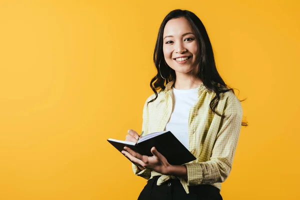 Happy asian girl writing in notebook with pen, isolated on yellow — Stock Photo