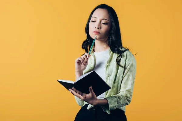 Nachdenkliches asiatisches Mädchen mit Notizbuch und Stift, isoliert auf gelb — Stockfoto
