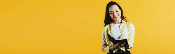Smiling asian girl writing in notebook with pen, isolated on yellow — Stock Photo