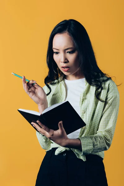 Conmocionado chica asiática escribiendo en cuaderno con pluma, aislado en amarillo - foto de stock