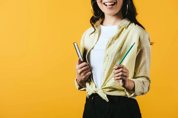 Обрезанный вид улыбающейся девушки с ноутбуком и ручкой, изолированный на желтый — стоковое фото