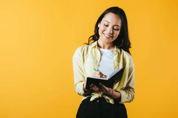 Attractive asian girl writing in notebook with pen, isolated on yellow — Stock Photo