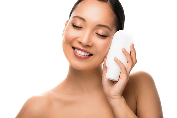 Усміхнена азіатська жінка з закритими очима тримає пляшку лосьйону, ізольована на білому — стокове фото