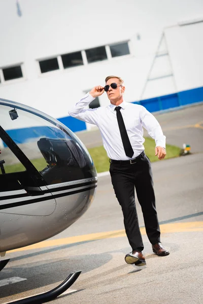 Piloto guapo en ropa formal ajustando gafas de sol y caminando cerca de helicóptero - foto de stock