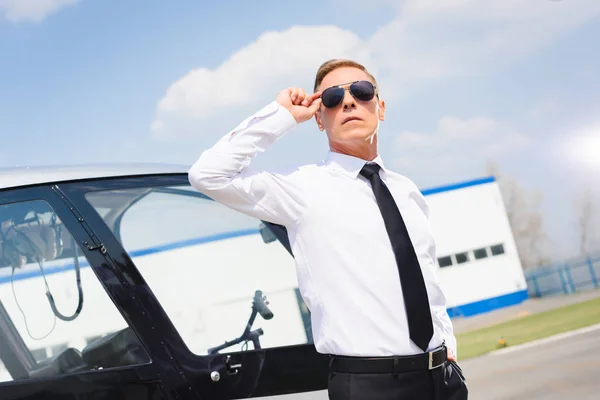 Piloto guapo en ropa formal ajustando gafas de sol cerca de helicóptero - foto de stock