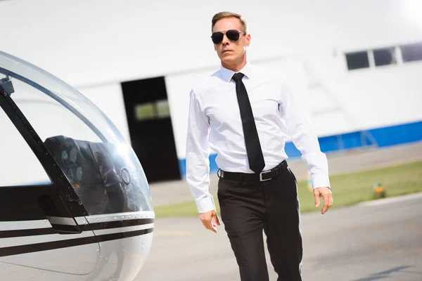 Piloto en ropa formal y gafas de sol caminando cerca de helicóptero - foto de stock