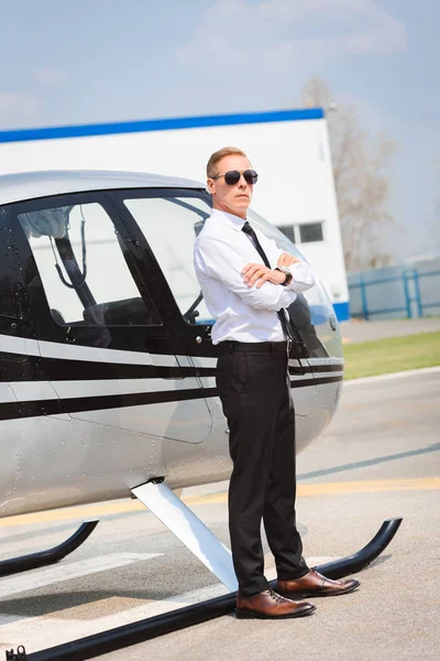 Pilot in sunglasses and formal wear with crossed arms posing near helicopter — Stock Photo