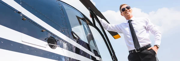 Prise de vue panoramique du pilote souriant en tenue formelle et lunettes de soleil près de l'hélicoptère — Photo de stock