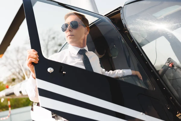 Reifer Pilot in offizieller Kleidung und Sonnenbrille öffnet Tür des Hubschraubers — Stockfoto