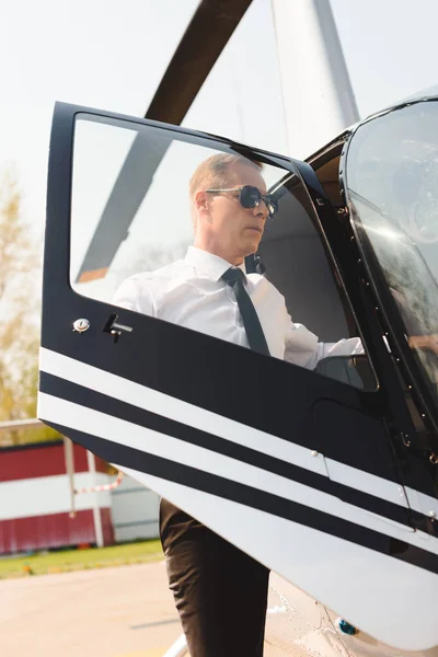 Piloto maduro en ropa formal y gafas de sol abriendo la puerta del helicóptero - foto de stock