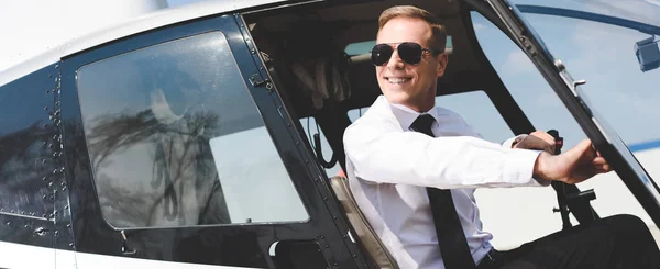 Plano panorámico de piloto sonriente guapo en ropa formal y gafas de sol en cabina de helicóptero - foto de stock