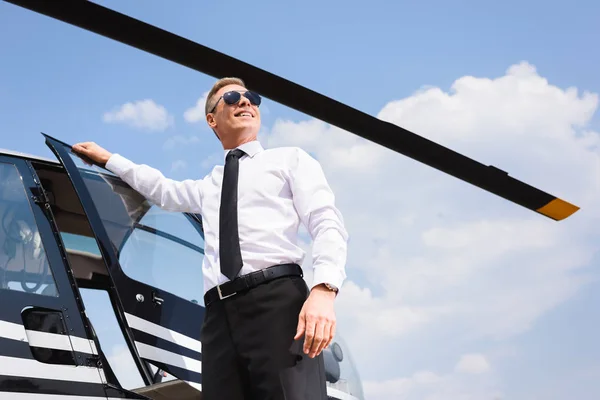 Vista de ángulo bajo de buen aspecto feliz piloto en desgaste formal y gafas de sol cerca de helicóptero - foto de stock