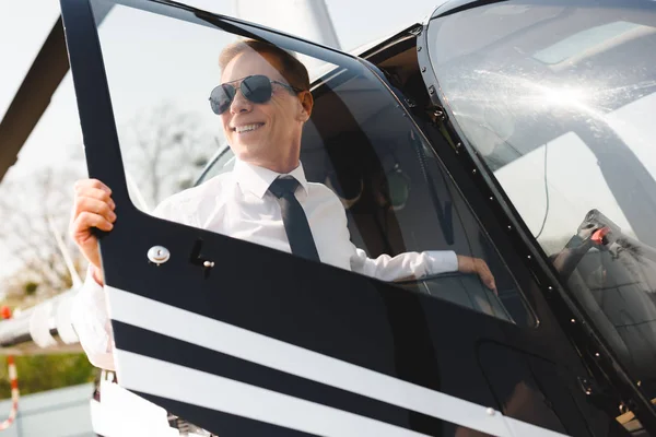 Sonriente madura Piloto en desgaste formal sentado abriendo la puerta de helicóptero - foto de stock