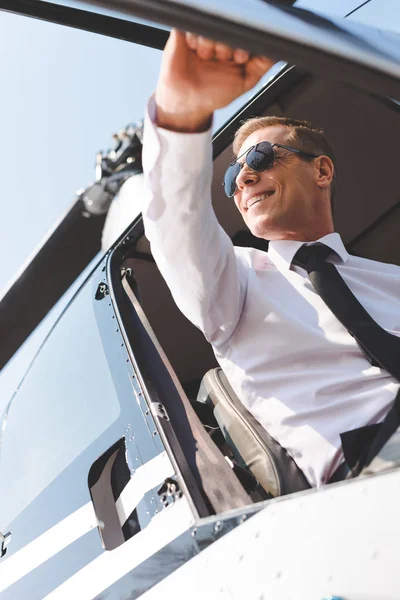 Smiling Pilot in sunglasses and formal wear sitting in helicopter cabin and opening door — Stock Photo
