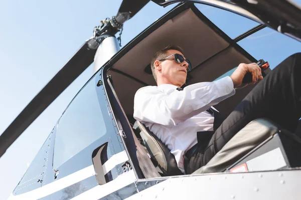 Piloto maduro en gafas de sol y ropa formal sentado en cabina de helicóptero - foto de stock