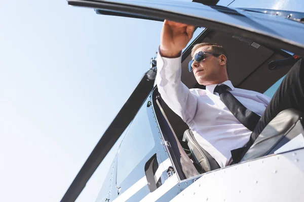 Piloto guapo en gafas de sol y ropa formal sentado en cabina de helicóptero y puerta de apertura - foto de stock