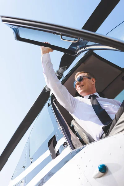 Vista de ángulo bajo de piloto en gafas de sol y desgaste formal sentado en cabina de helicóptero y puerta de apertura - foto de stock