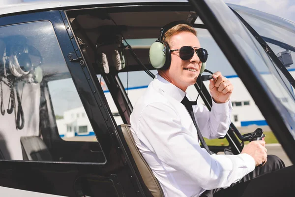 Piloto sonriente en gafas de sol y auriculares sentado en cabina de helicóptero - foto de stock