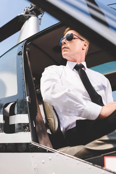 Vista de ángulo bajo de piloto guapo en gafas de sol y desgaste formal sentado en cabina de helicóptero - foto de stock