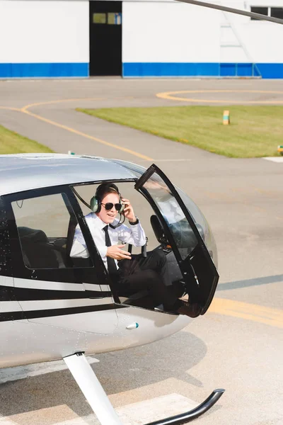 Piloto maduro en ropa formal y gafas de sol sentado en cabina de helicóptero con puerta abierta - foto de stock